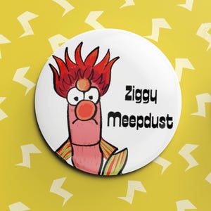 Peut inclure: Un badge blanc avec un personnage de dessin animé. Le personnage a des cheveux rouges, un nez rouge et de grands yeux. Le nom "Ziggy Meepdust" est écrit en noir. Le fond est jaune avec des éclairs blancs.