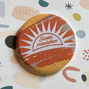Può includere: Un bottone rotondo con una grafica di alba e il testo "Sunrise, parabellum" in bianco su uno sfondo marrone, giallo e grigio.
