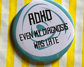 May include: A round blue and white button with the text "ADHD Even My Diagnosis Was Late" in black.