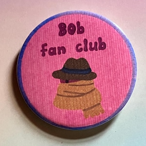 May include: A pink and blue round button with a cartoon illustration of a person wearing a brown hat and scarf. The text "Bob fan club" is written in pink letters on the button.