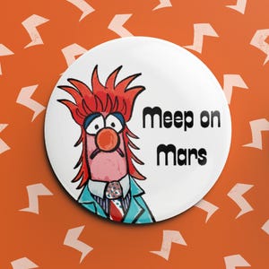 Peut inclure: Un badge blanc avec un personnage de dessin animé aux cheveux roux, un nez rouge et un costume bleu. Le texte "Meep on Mars" est imprimé en noir. Le fond est orange avec des éclairs blancs.