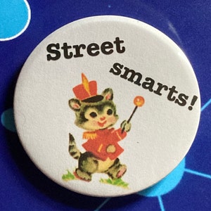 Può includere: Un bottone bianco con la scritta "Street Smarts!" in nero. Il bottone presenta un gatto dei cartoni animati in uniforme da banda rossa che tiene in mano una bacchetta. Il gatto indossa un cappello rosso con un pennacchio dorato.