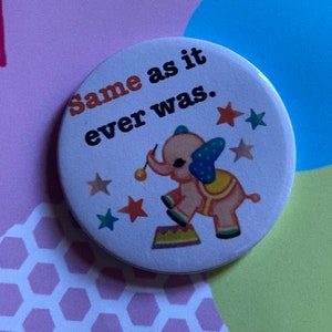 May include: A white button featuring a pink elephant balancing on a colourful circus ring. The text "Same as it ever was." is printed in black on the button.