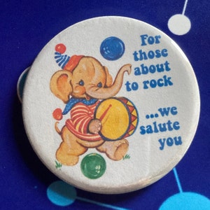 May include: A white button featuring a cartoon elephant playing a drum. The text on the button reads "For those about to rock...we salute you".