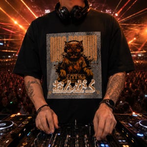 Puede incluir: Camiseta negra con un gráfico de un gato con auriculares y un equipo de DJ. La camiseta tiene texto japonés y las palabras "ELECTRONIC DANCE MUSIC". El fondo es una escena de concierto borrosa.