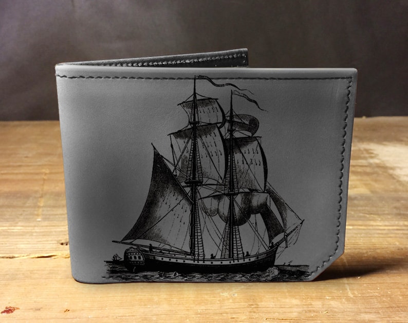 May include: A gray leather wallet with a black stitched border features an engraved image of a vintage sailing ship.