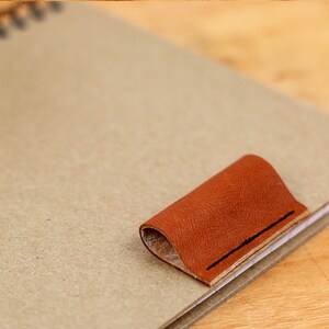 May include: A brown leather bookmark with a stitched edge is attached to a notebook.