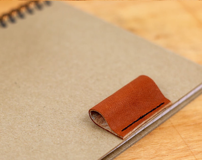 May include: A brown leather bookmark with a stitched edge is attached to a notebook.