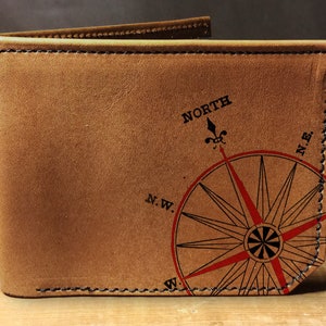 May include: A brown leather wallet with a compass rose design. The compass rose is red and black, with the word "NORTH" above it. The wallet is stitched with black thread.