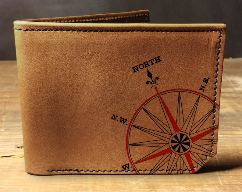 May include: A brown leather wallet with a compass rose design. The compass rose is red and black, with the word "NORTH" above it. The wallet is stitched with black thread.