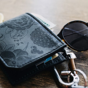 May include: A black leather wallet with a floral pattern, containing cash and cards, sits next to a pair of tortoiseshell sunglasses and a set of keys. The wallet is open, revealing its contents. The items are on a wooden surface.