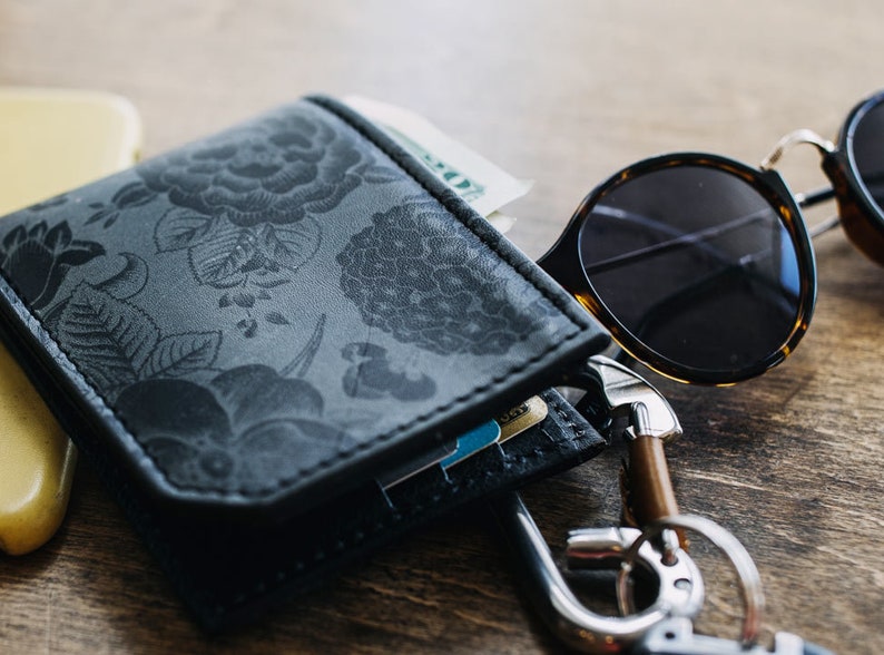 May include: A black leather wallet with a floral pattern, containing cash and cards, sits next to a pair of tortoiseshell sunglasses and a set of keys. The wallet is open, revealing its contents. The items are on a wooden surface.