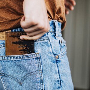 May include: A brown leather wallet with a black forest silhouette design is partially visible in the back pocket of blue jeans. The wallet is rectangular and appears to be made of textured leather.