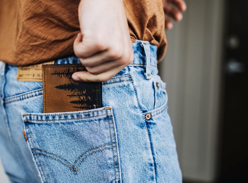 May include: A brown leather wallet with a black forest silhouette design is partially visible in the back pocket of blue jeans. The wallet is rectangular and appears to be made of textured leather.