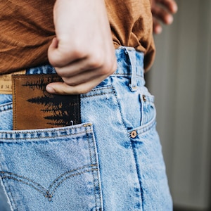 May include: A brown leather wallet with a black silhouette of trees printed on it. The wallet is being placed in the back pocket of a pair of blue jeans.