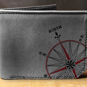May include: Gray leather wallet with a compass rose design. The compass rose is red and black, with the word "NORTH" above it. The wallet is folded open, showing the interior.