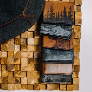 May include: A stack of eight leather wallets with different designs. The wallets are arranged vertically, with the top wallet featuring a forest scene and the bottom wallet featuring a floral design. The wallets are all brown or black and have a stitched edge.