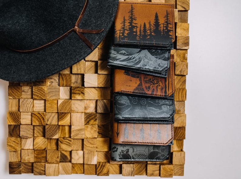 May include: A stack of eight leather wallets with different designs. The wallets are arranged vertically, with the top wallet featuring a forest scene and the bottom wallet featuring a floral design. The wallets are all brown or black and have a stitched edge.