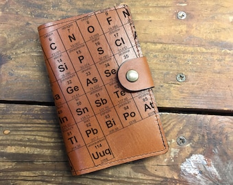 May include: A brown leather notebook with a periodic table design. The notebook has a flap closure with a metal button.