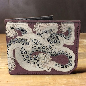 Peut inclure: Un portefeuille bifold violet avec une illustration détaillée d'un léopard des neiges. Le léopard est blanc avec des taches noires, et le portefeuille a un intérieur et des coutures noirs.