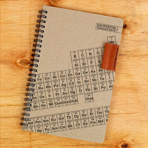May include: A brown notebook with a spiral binding and a periodic table printed on the cover. The notebook has a brown leather bookmark attached to the top.