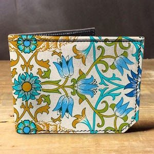 May include: A floral patterned wallet with a design of blue and teal flowers with gold and green accents on a cream background. The wallet has a black interior and is made of leather.