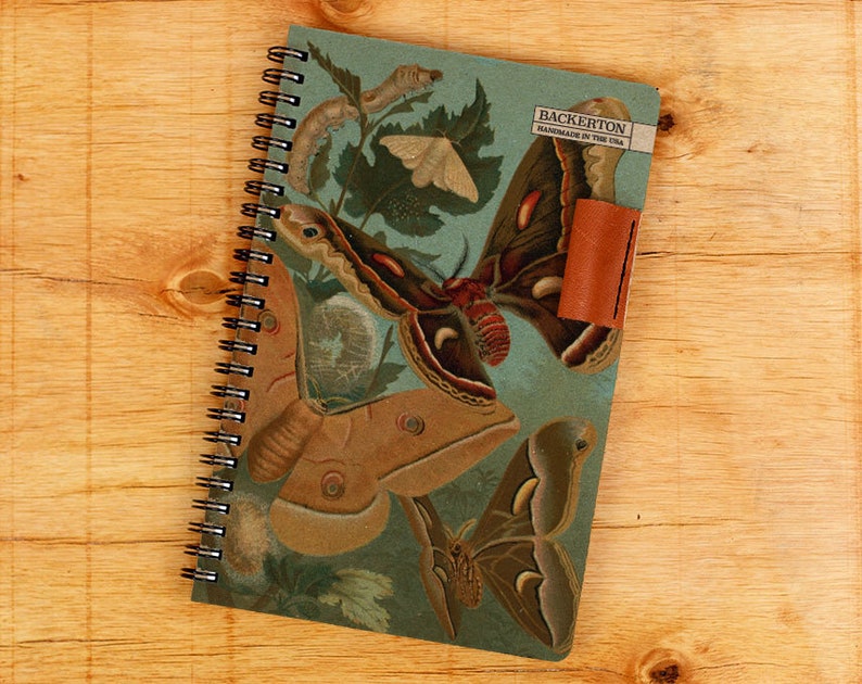 May include: A blue spiral bound notebook with a cover featuring a vintage illustration of moths, butterflies, and caterpillars. The cover also includes the text "Backerton Handmade in the USA".