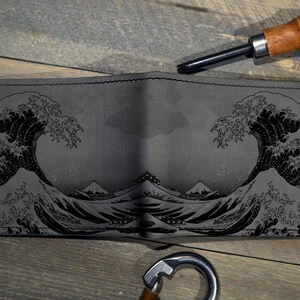 Op de afbeelding: Grijze leren portemonnee met een zwart reli&euml;f ontwerp van de Grote Golf van Kanagawa. De portemonnee is open en toont de binnenkant. Een leerbewerkingstool en een metalen karabijnhaak zijn ook zichtbaar.