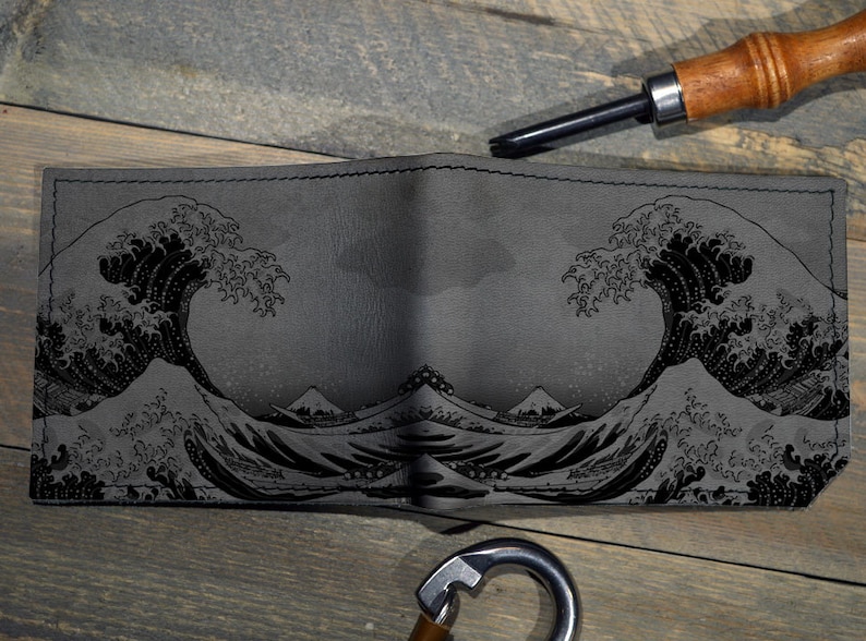 Op de afbeelding: Grijze leren portemonnee met een zwart reli&euml;f ontwerp van de Grote Golf van Kanagawa. De portemonnee is open en toont de binnenkant. Een leerbewerkingstool en een metalen karabijnhaak zijn ook zichtbaar.