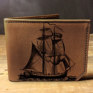 May include: A brown leather wallet with an engraved image of a sailing ship. The ship has multiple sails and is detailed with ropes and rigging.