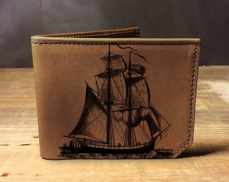 May include: A brown leather wallet with an engraved image of a sailing ship. The ship has multiple sails and is detailed with ropes and rigging.