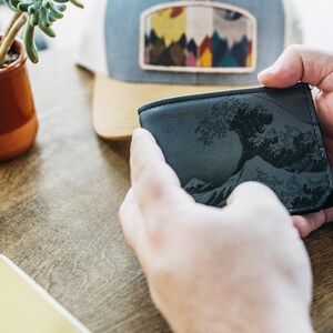 May include: A black leather wallet featuring a detailed wave design. The wallet is held in two hands, showcasing the intricate artwork. A cap with a mountain scene patch is in the background.