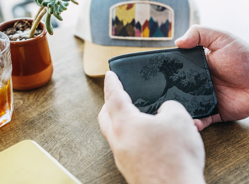 May include: A black leather wallet featuring a detailed wave design. The wallet is held in two hands, showcasing the intricate artwork. A cap with a mountain scene patch is in the background.