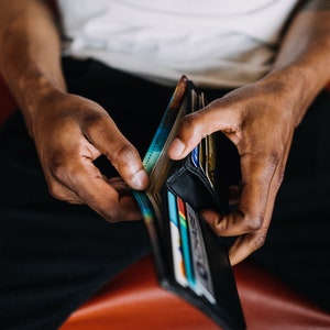 May include: A black leather wallet is open, revealing multiple credit cards and cash. The wallet is held by a person wearing black pants. The background is a blurred red surface.