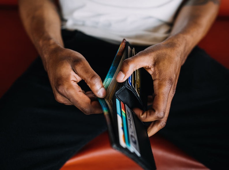 May include: A black leather wallet is open, revealing multiple credit cards and cash. The wallet is held by a person wearing black pants. The background is a blurred red surface.