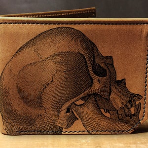 May include: A tan leather wallet with a detailed black illustration of a human skull on the front. The wallet is open, revealing the interior. The edges are stitched with black thread, and the leather has a slightly distressed appearance.