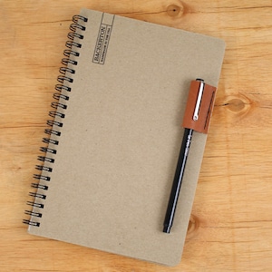May include: A brown notebook with a black spiral binding and a brown leather pen holder. The notebook has the text "BACKERTON HANDMADE IN THE USA" printed on the cover.