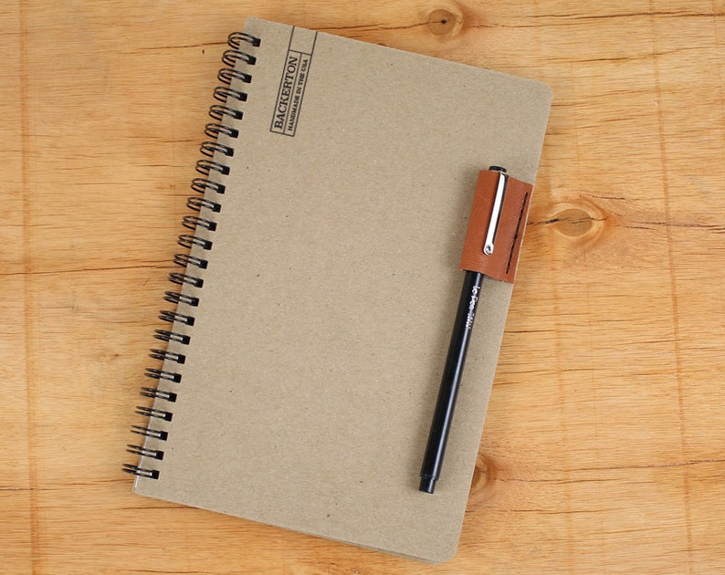May include: A brown notebook with a black spiral binding and a brown leather pen holder. The notebook has the text "BACKERTON HANDMADE IN THE USA" printed on the cover.