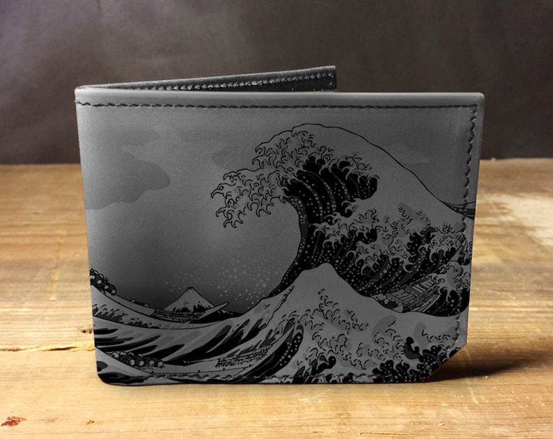 Op de afbeelding: Een grijze bi-fold portemonnee met een zwart-wit illustratie van een grote oceaangolf en een berg. De portemonnee is gemaakt van een glad materiaal en heeft een gestikte rand.