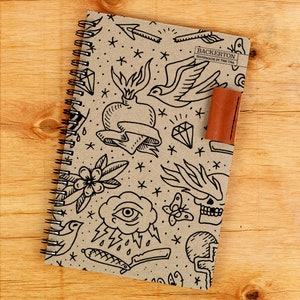 May include: A brown notebook with a spiral binding and a leather strap. The cover features a black and white illustration of various traditional tattoo designs, including a heart, a bird, a skull, and a diamond. The text "Backerton Handmade in the USA" is printed on the cover.