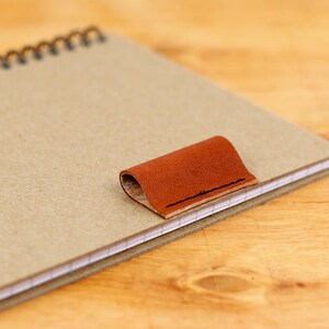 May include: A brown leather bookmark attached to a spiral bound notebook with beige paper.