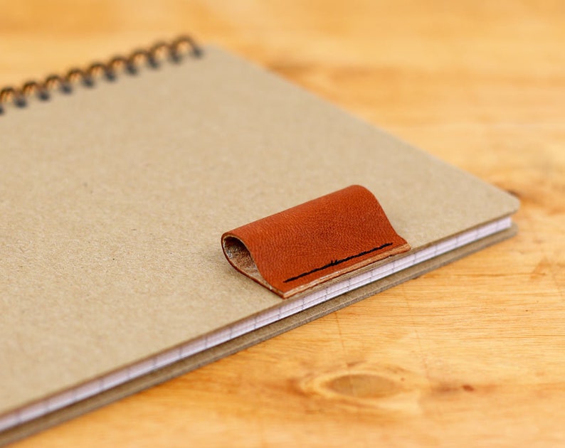 May include: A brown leather bookmark attached to a spiral bound notebook with beige paper.
