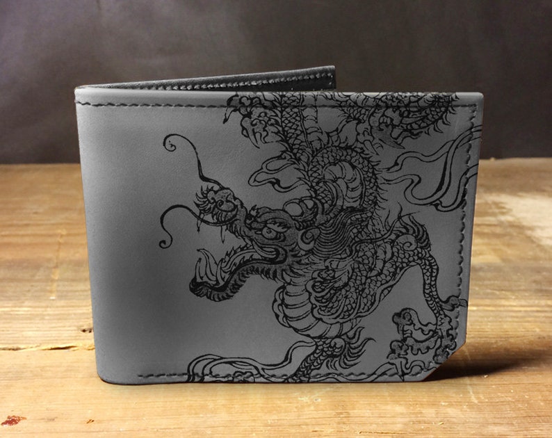 Puede incluir: Una cartera de cuero gris con un dise&ntilde;o de drag&oacute;n negro. El drag&oacute;n est&aacute; detallado con escamas y espirales intrincados.