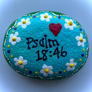 May include: Hand-painted, oval-shaped stone with a turquoise background. White daisies with yellow centers and purple and yellow dots surround the edges. The text "Psalm 18:46" is written in black, and a red heart is painted on the stone.