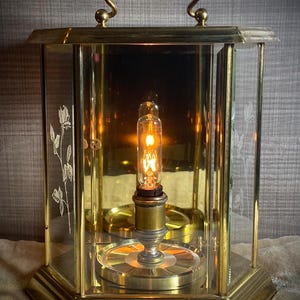 May include: A gold-toned, octagonal, glass-enclosed clock with a clear glass face and a single light bulb inside. The clock has a decorative handle on the top and etched floral designs on the sides.