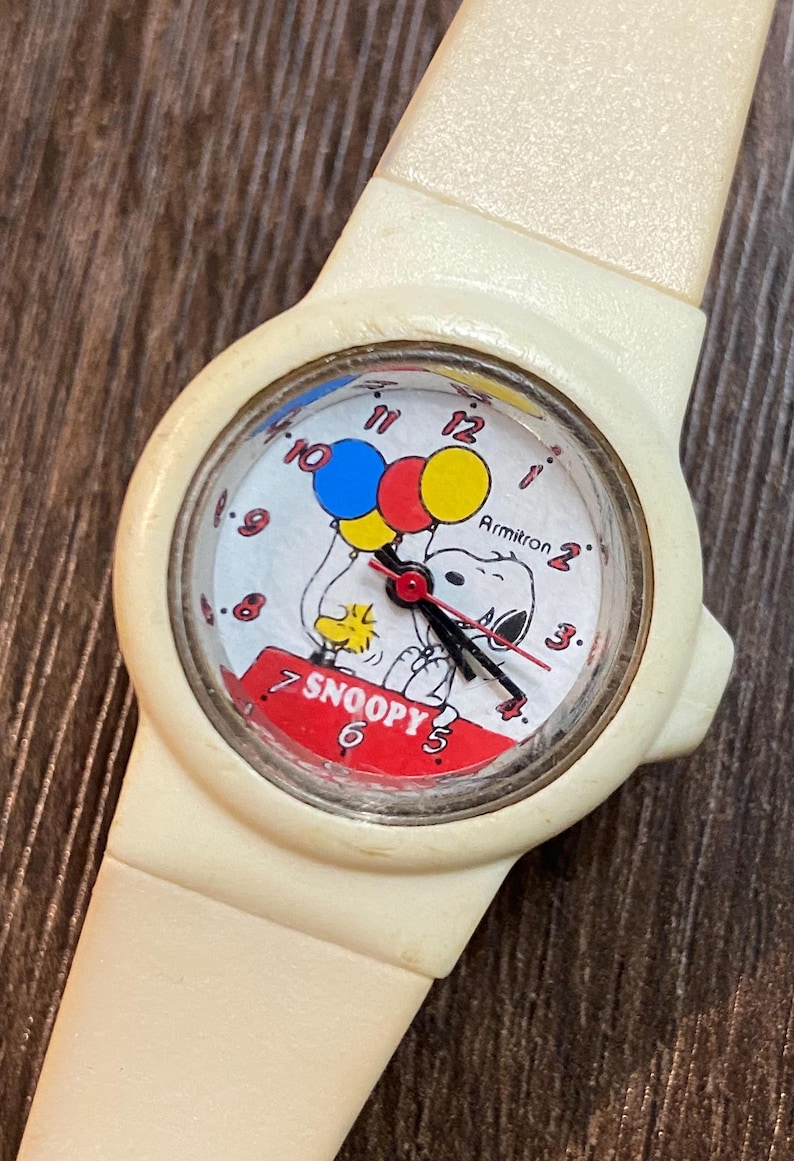 May include: Cream-colored Snoopy wristwatch with a round face. The watch face features Snoopy holding balloons and the word "Snoopy". The watch has black hands and a cream-colored band.