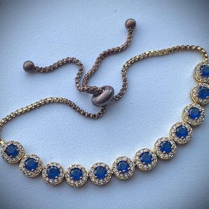 May include: A gold-toned bracelet featuring a series of round, blue gemstones encircled by smaller, clear stones. The bracelet has a delicate chain and an adjustable sliding closure. The gemstones are evenly spaced along the bracelet's length.