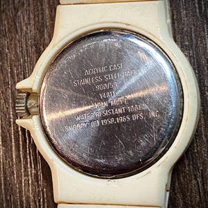 May include: Close-up of a vintage cream-colored Snoopy watch. The watch has a round face with a stainless steel back. The text on the back reads "ACRYLIC CASE, STAINLESS STEEL BACK, WATER RESISTANT 100FT, SNOOPY (C) 1958.1965 UFS, INC."