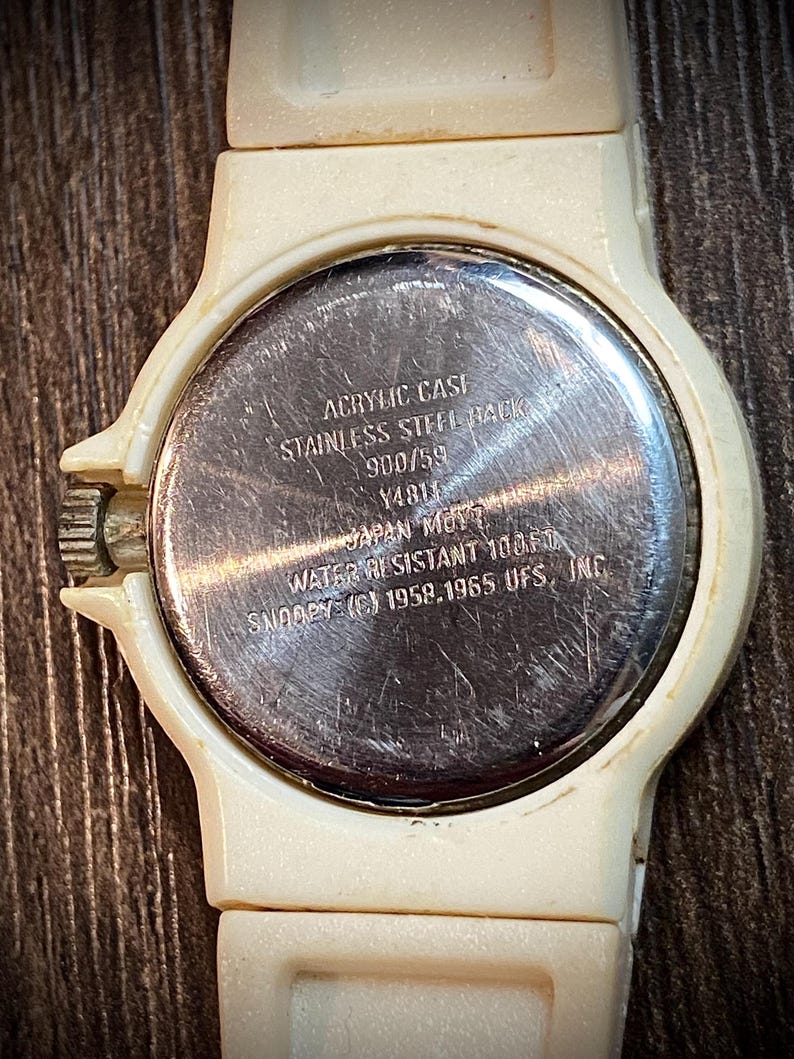 May include: Close-up of a vintage cream-colored Snoopy watch. The watch has a round face with a stainless steel back. The text on the back reads "ACRYLIC CASE, STAINLESS STEEL BACK, WATER RESISTANT 100FT, SNOOPY (C) 1958.1965 UFS, INC."