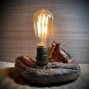 May include: A rustic wooden lamp base with a warm, glowing Edison bulb. The base is made from a piece of driftwood with a natural, weathered finish.